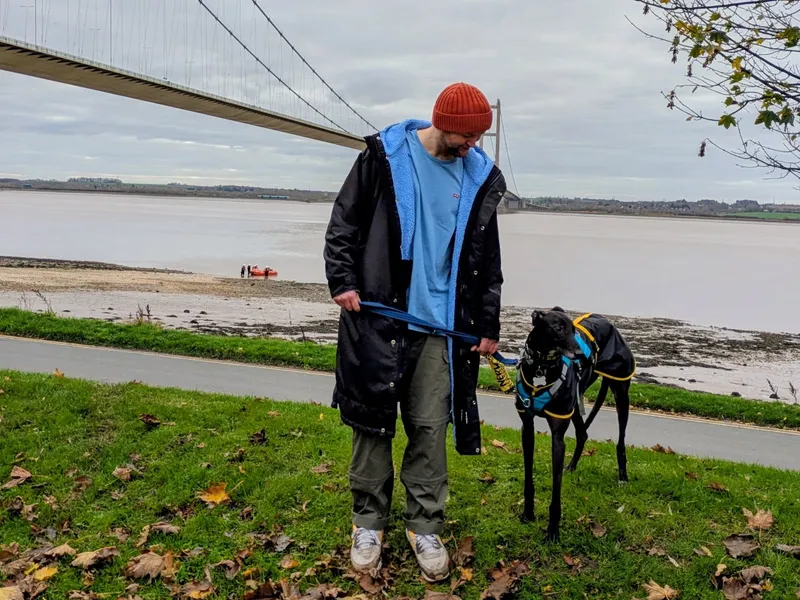 A man wearing an orange beanie and black raincoat stands next to his black greyhound and gazes at him lovingly. The greyhound is looking off-camera. There is a bridge and river in the background.